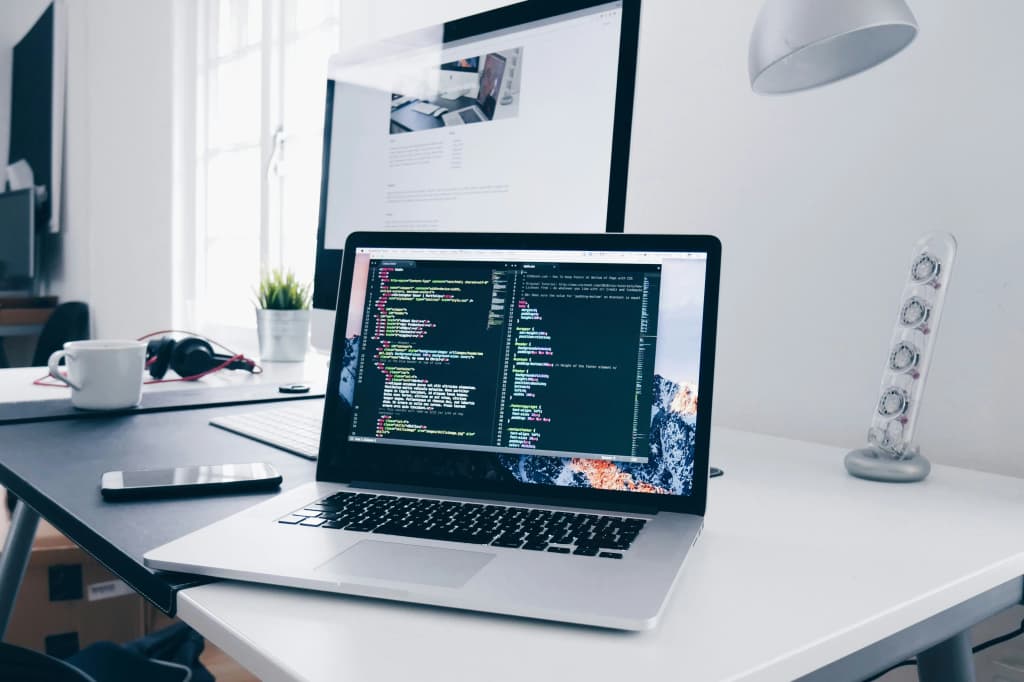 Laptop displaying source code in a dark editor on a white desk, with a monitor, headphones, coffee mug, and phone nearby