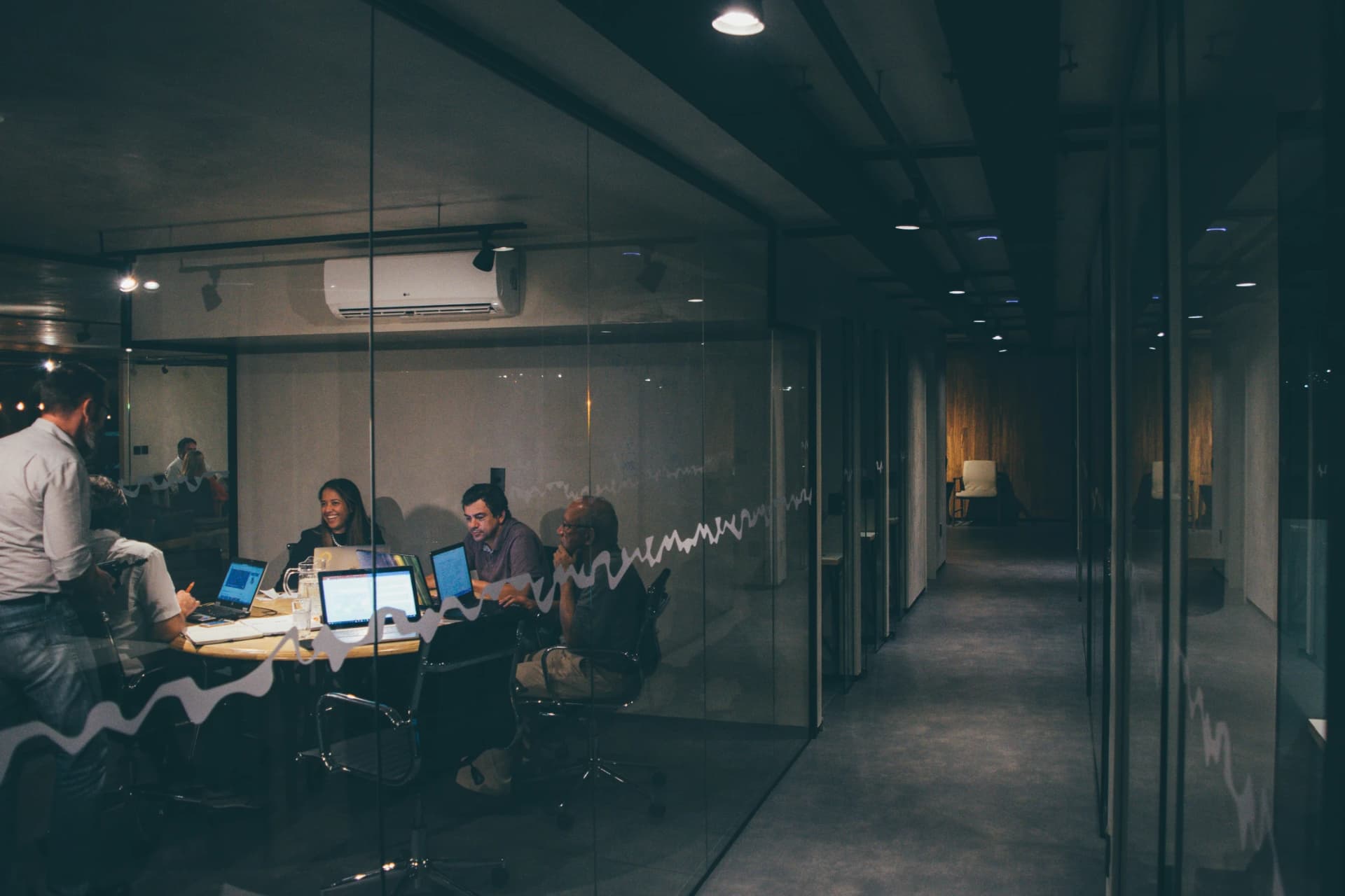 Team meeting at night in a glass-walled office conference room, with people around a table with laptops beside a dim corridor