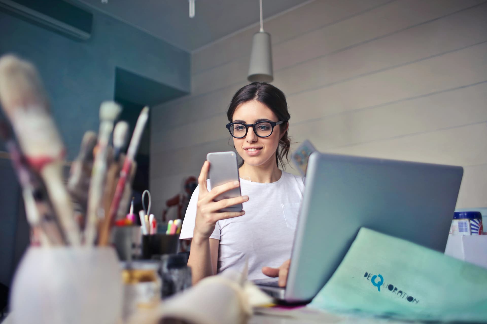 Smiling woman in glasses looking at her phone while sitting at a laptop in a bright workspace with art supplies in the foreground