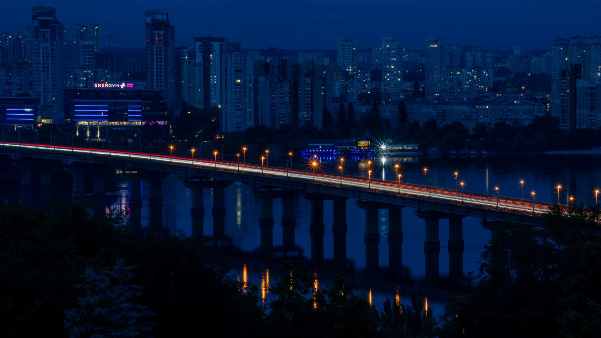 Long illuminated bridge crossing a river at dusk in front of a city skyline of lit high-rise buildings