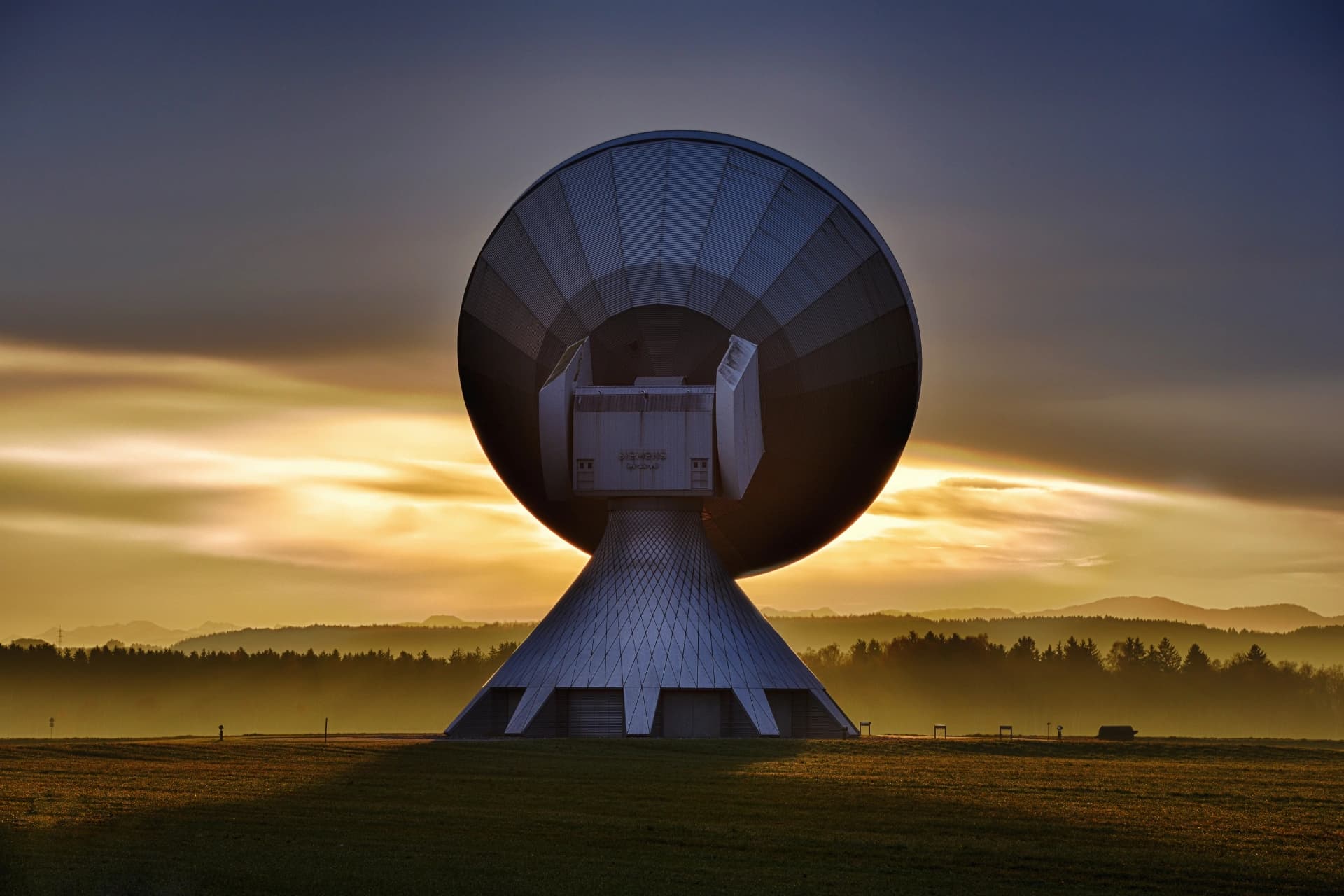 A large satellite dish antenna on a conical base in a field, silhouetted against a golden sunset sky with forested hills behind it