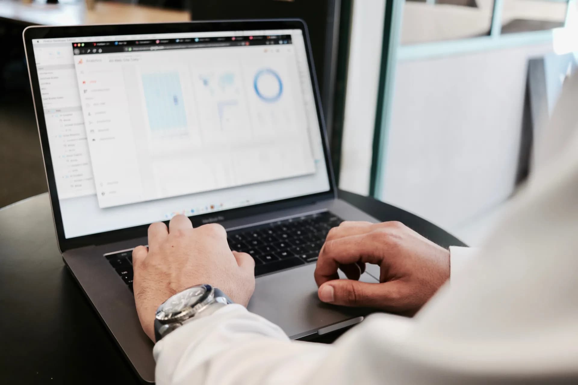 Over-the-shoulder view of a person wearing a wristwatch typing on a laptop that displays an analytics dashboard with charts and a world map