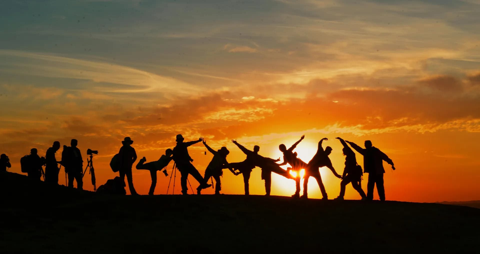 Silhouettes of a group of people striking playful balancing poses on a hilltop against a bright orange sunset, with photographers and tripods at the left