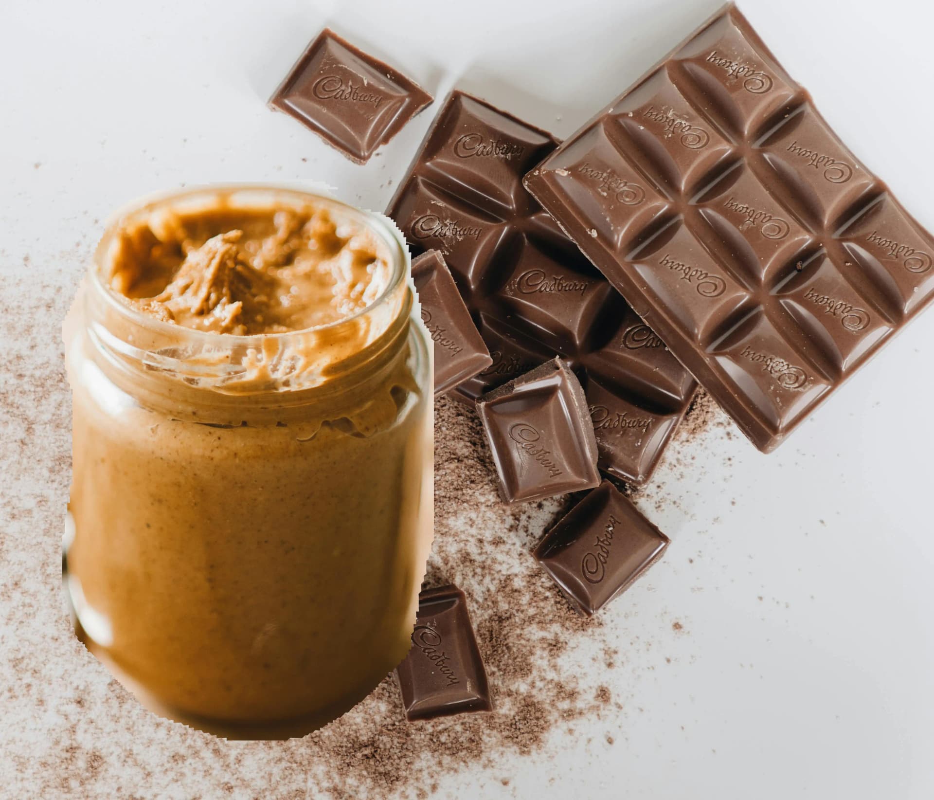 Open jar of peanut butter beside broken squares of Cadbury milk chocolate scattered with cocoa powder on a white surface