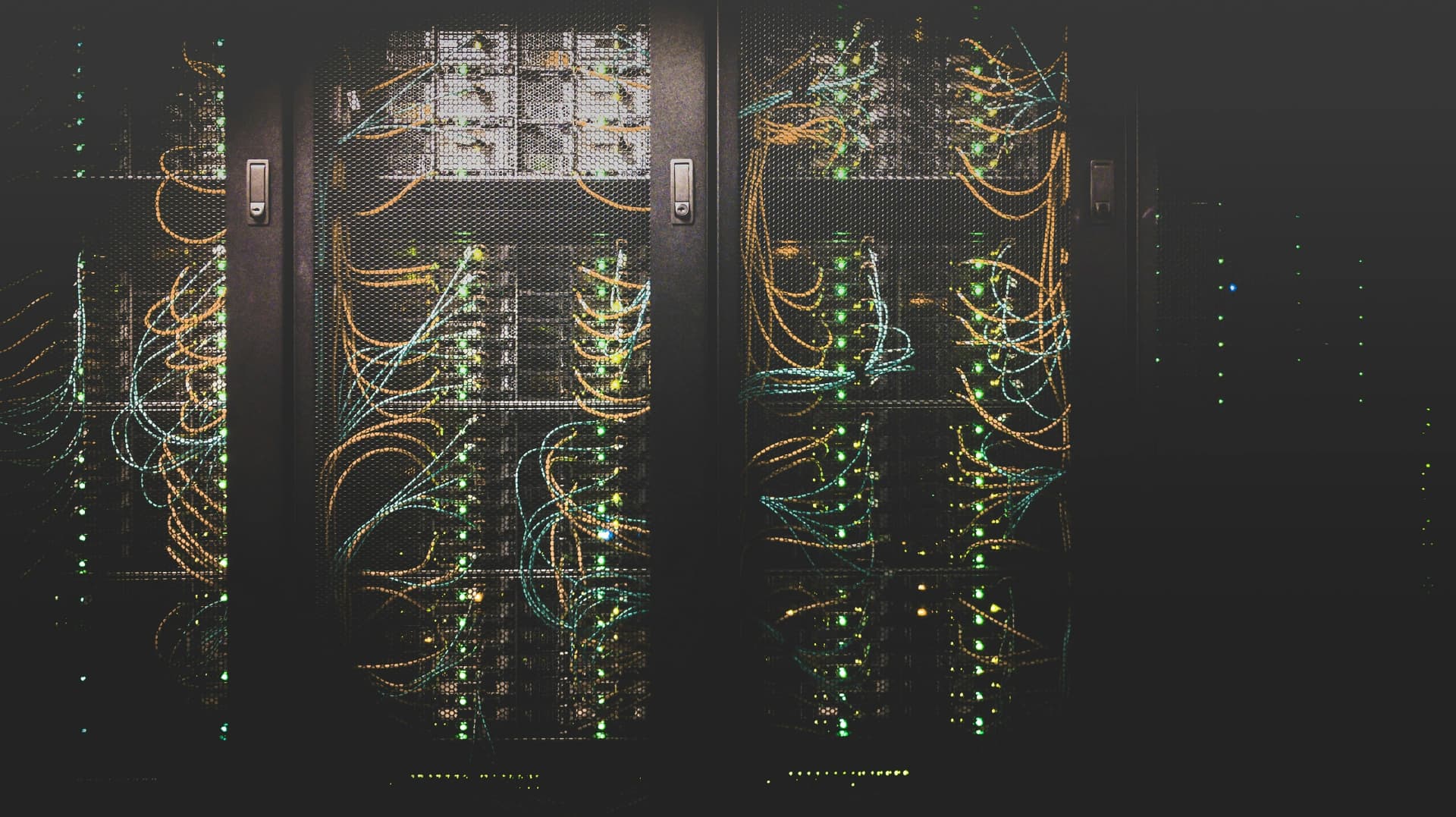 Dark server racks behind mesh doors, filled with tangles of orange and teal network cables and green status lights