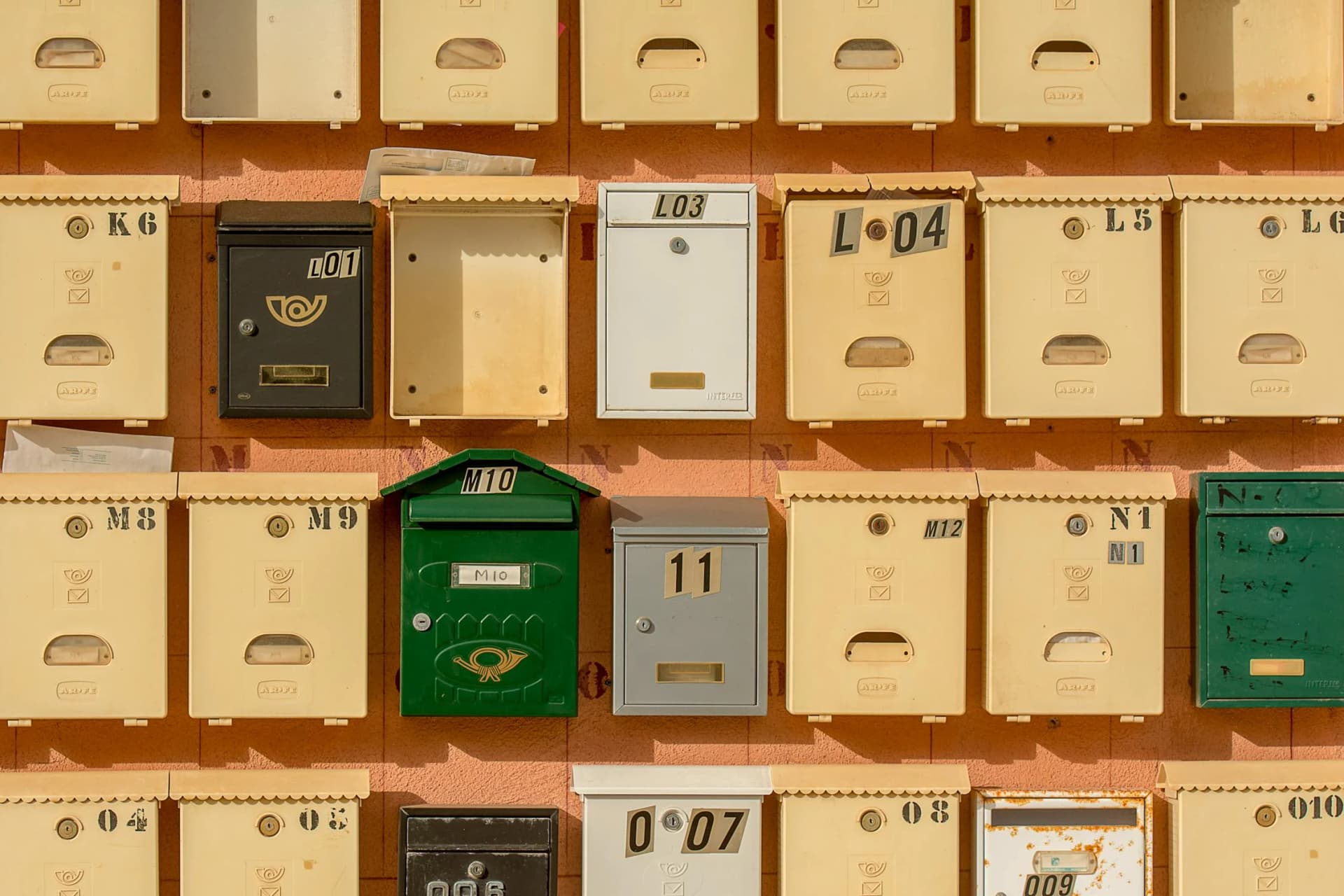 Wall of numbered mailboxes, mostly cream with a few black, green, white, and gray ones, labeled with codes like L01, M10, and N1