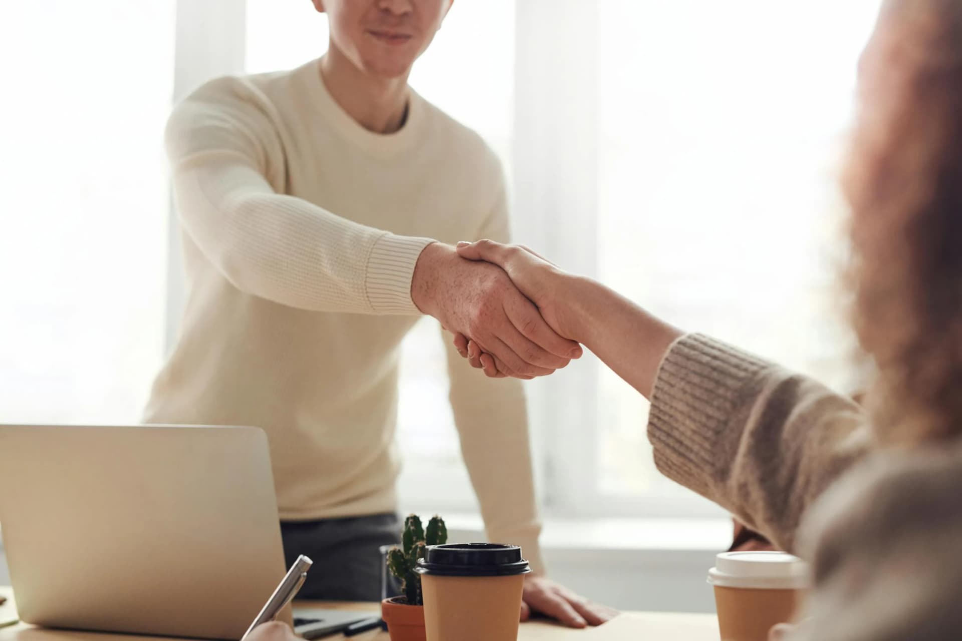 Close-up of two people shaking hands across a table with a laptop, takeaway coffee cups, and a small cactus