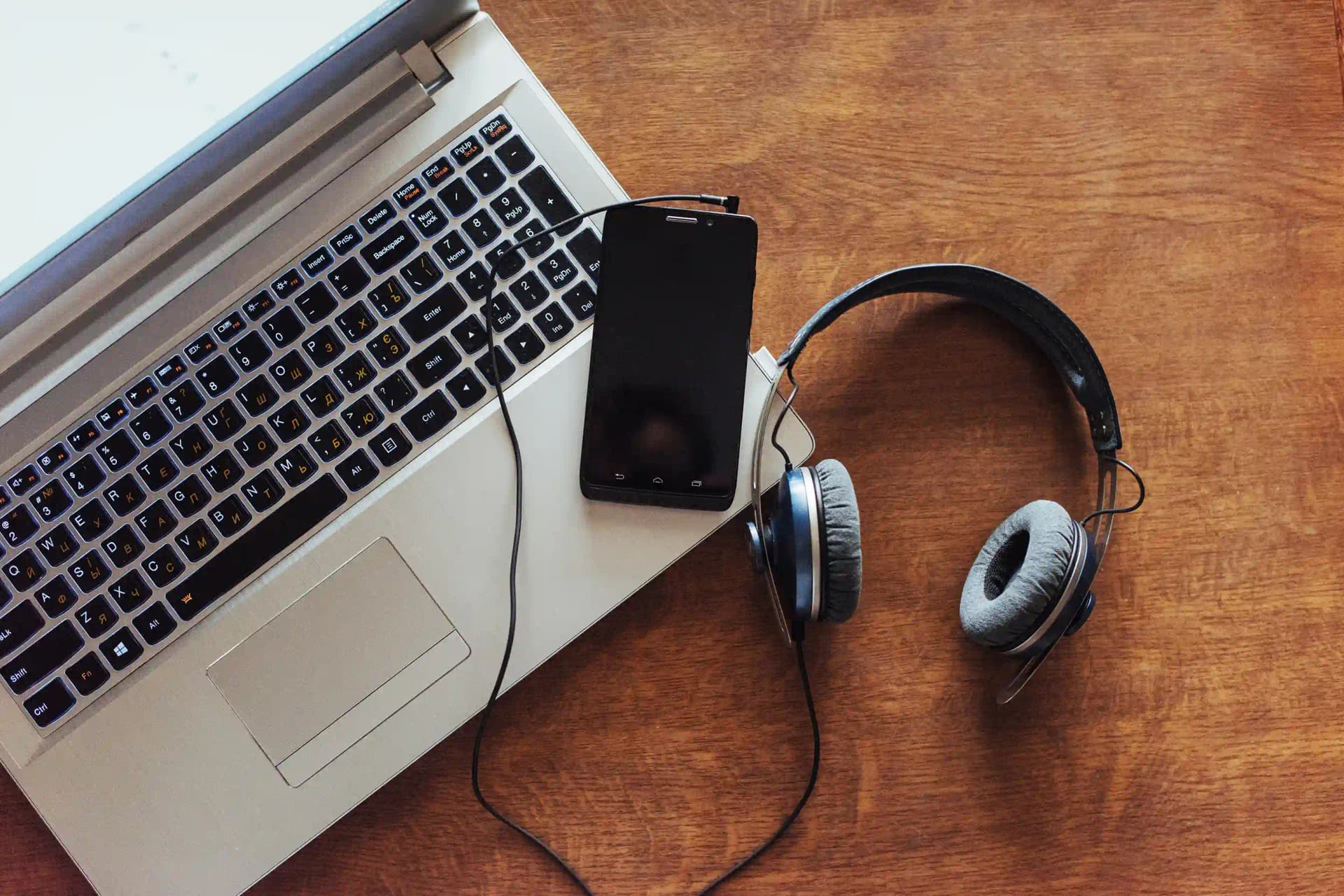 Overhead view of a laptop keyboard on a wooden desk with a smartphone resting on it and a pair of wired headphones beside it